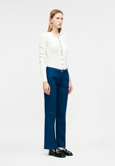 Γυναίκα που στέκεται, φορώντας μια λευκή ζακέτα με κουμπιά και μακριά μανίκια, μπλε παντελόνι με φαρδιά μπατζάκια και μαύρα loafers, μπροστά από ένα μονόχρωμο φόντο.