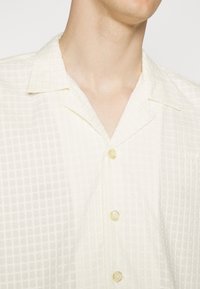 Crème-kleurig shirt met korte mouwen, een gestructureerd ruitpatroon, een notch-kraag en doorzichtige knoopsluitingen. Subtiele geborduurde logo-details.
