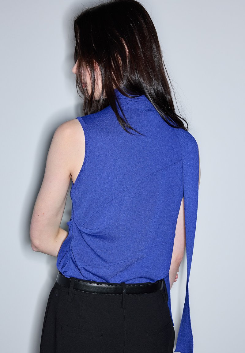 Mujer con cabello largo y oscuro lleva una blusa sin mangas azul con un detalle largo de tela en la espalda, metida dentro de pantalones negros con cinturón.