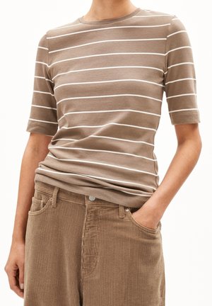 Persoon die een bruin-wit gestreept kortermig shirt draagt, ingestopt in een bruine corduroy broek met één hand in de zak.