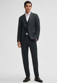 Dunkelgrüner Anzug bestehend aus einem maßgeschneiderten Jacket und einer Weste, kombiniert mit einem hellen Hemd und schmal geschnittenen Hosen. Schwarze Lederschuhe runden das Outfit ab.