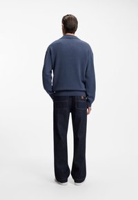 Blue knit sweater with a ribbed collar, paired with dark denim jeans featuring back pockets. Model stands with a neutral background.