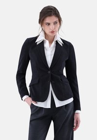 Marineblaue Cord-Blazer mit taillierter Passform, Reverskragen, zwei vorderen Taschen und einfachem Knopfverschluss, kombiniert mit einem weißen Hemd mit Kragen.