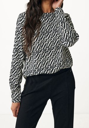 Vrouw draagt een zwart-wit blouse met geometrisch patroon en lange mouwen, ingestopt in hoge zwarte broeken, poseert met hand dicht bij haar gezicht.