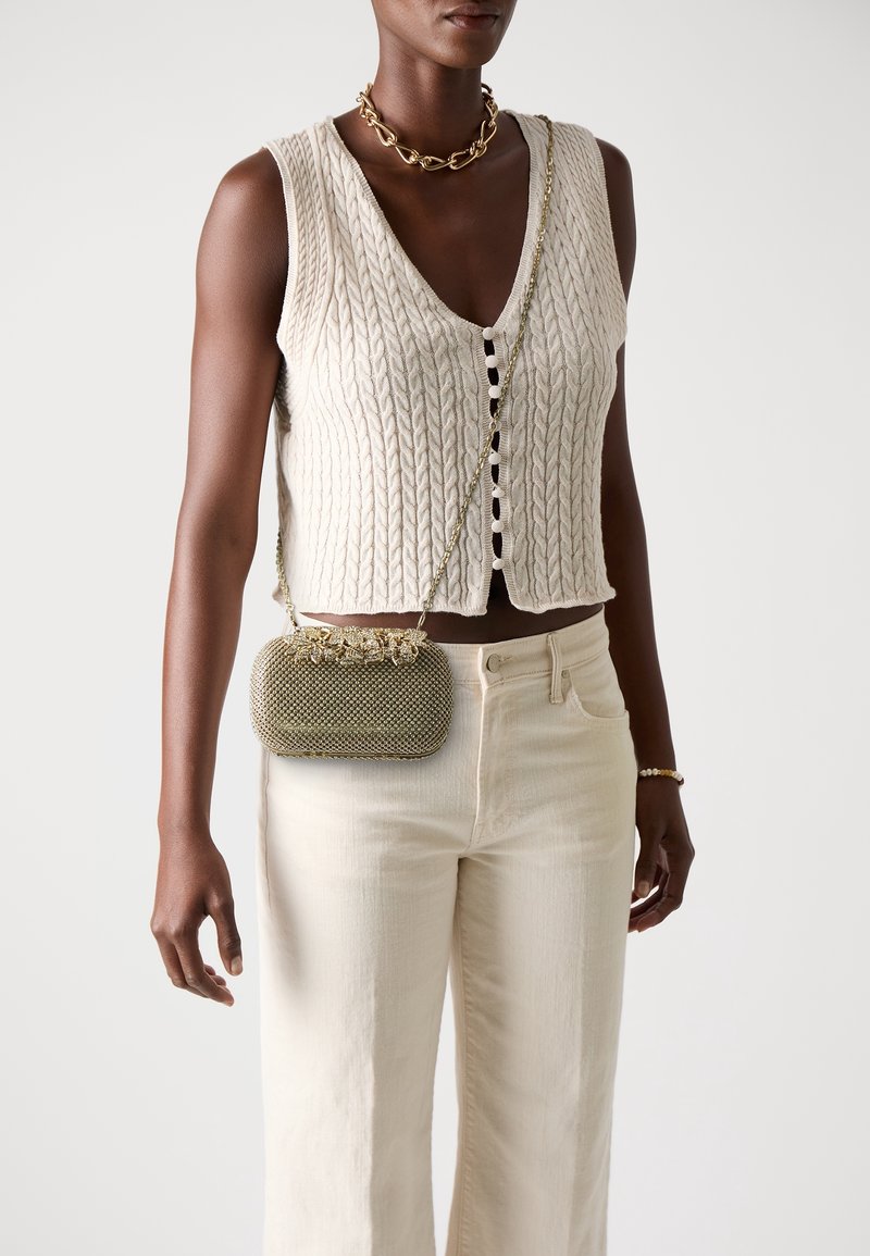 Femme portant un gilet sans manches en maille torsadée crème, un pantalon crème, un collier chaîne doré, un bracelet et un petit sac bandoulière doré texturé.