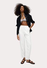 Black button-up shirt, striped halter bikini, white drawstring pants, and black sandals. Model poses with hands in pockets, neutral background.