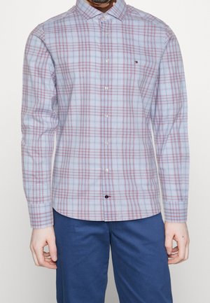 Male model wearing a light blue and red plaid button-up shirt with long sleeves and blue pants, standing against a plain background.