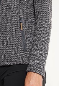 Giacca grigia con trama, dotata di una tasca con zip, caratterizzata da un motivo intrecciato e tessuto elasticizzato. Primo piano sul dettaglio della manica e della tasca.