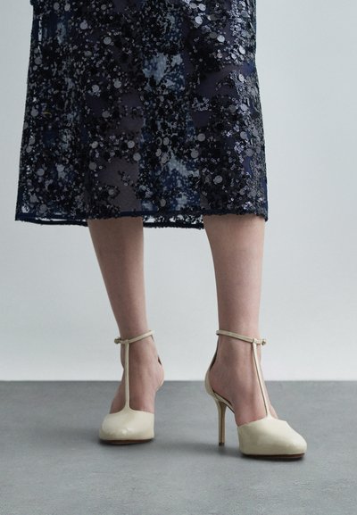 Beige patent leather high-heeled shoes with an open back, T-strap design, and a smooth texture.