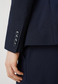 Μπλέιζερ σε ναυτικό χρώμα με μανίκι που διαθέτει τέσσερις στρογγυλές μαύρες κουμπότρυπες. Το ύφασμα έχει υφασμένη επιφάνεια, προσφέροντας κομψή εφαρμογή με δομημένο σχέδιο.