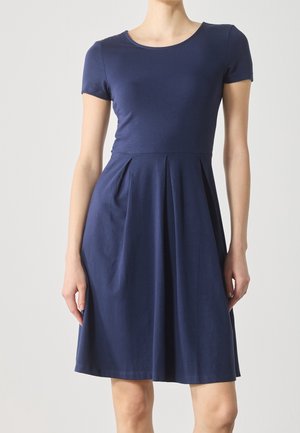 Donna che indossa un abito blu navy a maniche corte con un corpetto attillato e una gonna plissettata fino al ginocchio, in piedi su uno sfondo chiaro e semplice.