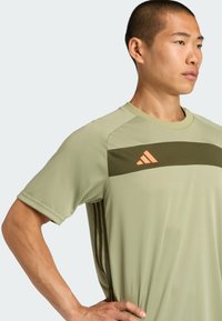 Camiseta atlética de color verde claro con una franja horizontal de color verde oscuro y un logo naranja. Presenta mangas cortas y una tela suave y texturizada.