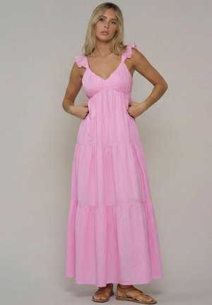 Blondine met een mouwloze roze maxi-jurk met geplooide bandjes en bruine platte sandalen met bandjes, staand tegen een effen achtergrond.