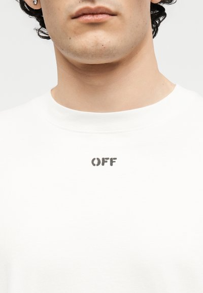 Κοντινή λήψη ενός ατόμου που φοράει λευκό πουκάμισο με στρογγυλή λαιμόκοψη, με τη λέξη "OFF" τυπωμένη με μικρά μαύρα γράμματα στο στήθος.