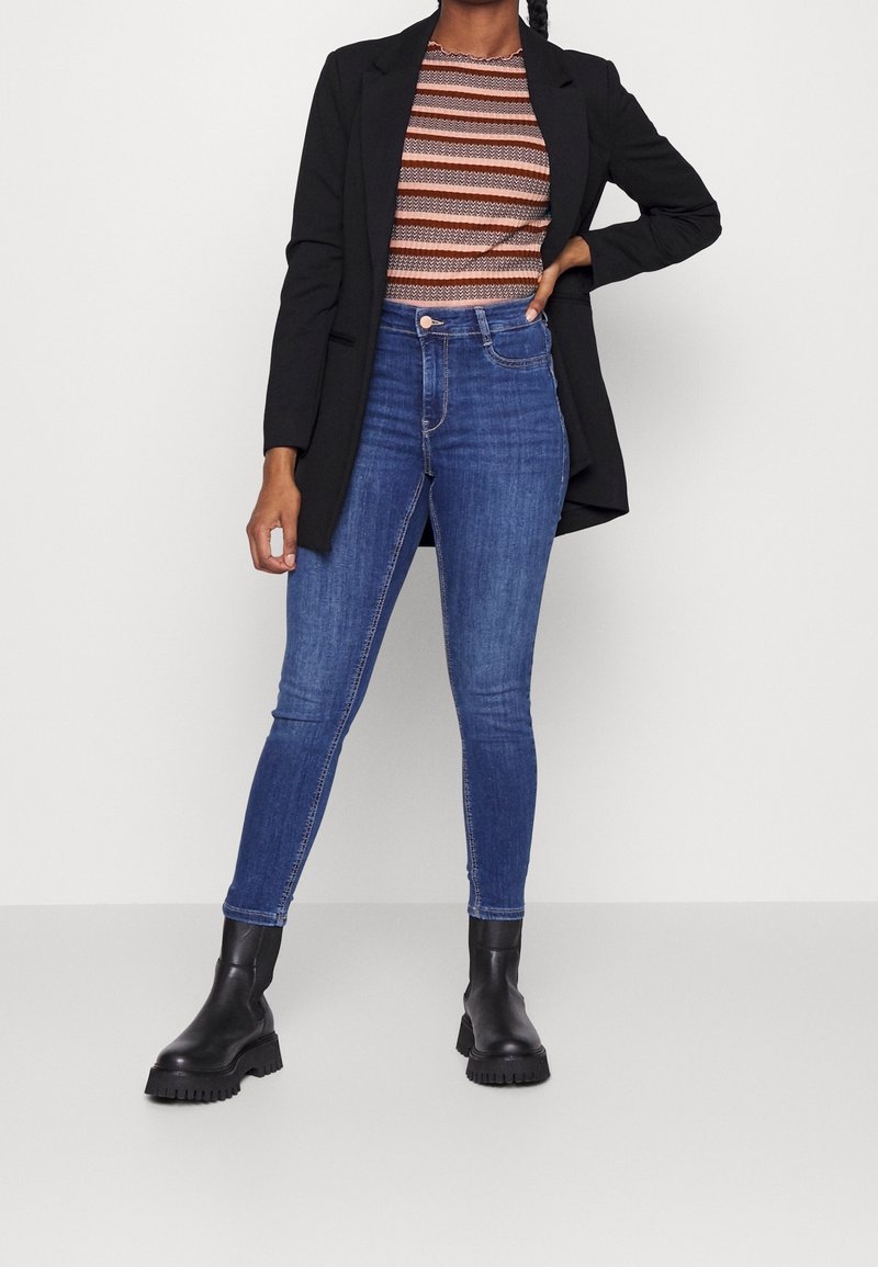 Sort blazer over en stribet brun og orange top, parret med blå skinny jeans og sorte chunky støvler, med struktureret stof og tætsiddende design.