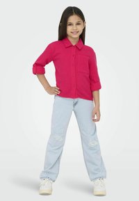 Camisa fucsia de botones con mangas enrolladas, bolsillo frontal, combinada con jeans claros rasgados y zapatillas blancas. Un atuendo sencillo y casual.