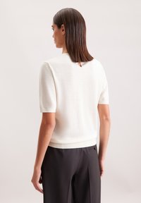 Kurzärmeliges, cremefarbenes Strickoberteil mit rundem Ausschnitt und kleinem Schlüsselloch hinten, kombiniert mit schwarzen Tailor-Pants.
