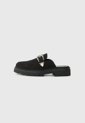 Czarne zamszowe buty wsuwane z grubą czarną gumową podeszwą i srebrnym zapięciem na cholewie. Gładka tekstura z zaokrąglonym kształtem.