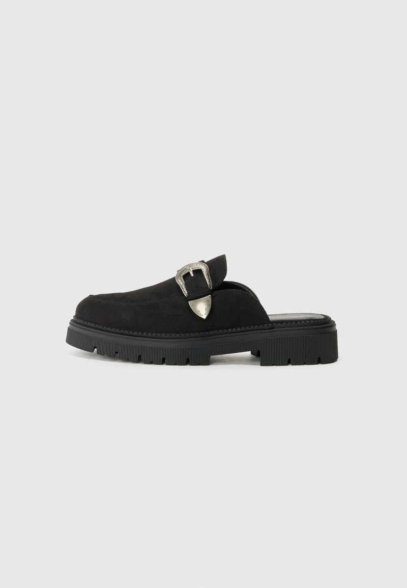 Sorte semskede slip-on sko med en tykk svart gummisåle og en sølvspenne-detalj på oversiden. Glatt tekstur med rund form.