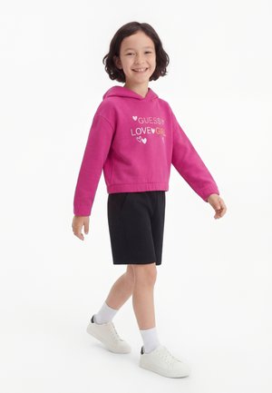 Χαμογελαστό παιδί που περπατά, φορώντας μια φωτεινή ροζ μπλούζα με κουκούλα με το κείμενο "Guess Love Girl", μαύρο σορτς, άσπρες κάλτσες και άσπρα αθλητικά παπούτσια σε λευκό φόντο.