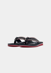 Un par de chanclas de Tommy Hilfiger en azul marino, rojo y blanco, con tiras de marca y suela roja, exhibidas sobre un fondo blanco.