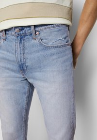 Světle modré džíny z denimu s jemným vyblednutím, opotřebované detaily na stehně, zapínání na knoflíky a klasický pětikapsový design.