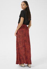 Rode maxi-rok met een abstract zwart patroon, voorzien van een hoge taille en vloeiende stijl. Gecombineerd met een zwart top met korte mouwen en beige schoenen.