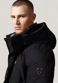 Czarna izolowana kurtka z wysokim kołnierzem i kapturem ściąganym na sznurki. Posiada subtelne logo na rękawie oraz zamek z przodu. Gładka faktura.