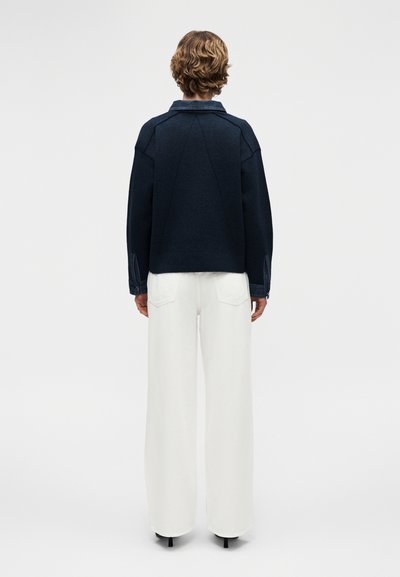 Personne aux cheveux courts et bouclés portant une veste bleu marine foncé et un pantalon blanc à jambes larges, debout de dos sur un fond uni.