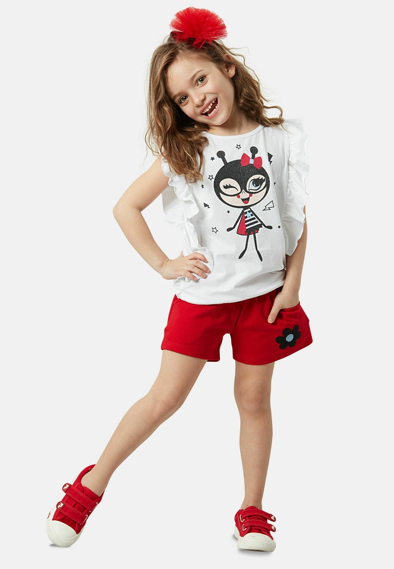 Camiseta blanca con mangas acampanadas y gráfico de una chica de dibujos animados, combinada con pantalones cortos rojos con un parche de flor y zapatillas rojas.