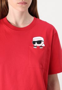 Raudona medvilninė marškinėliai su apvalia iškirpte, su cartoon grafika, vaizduojančia figūrą su akinių nuo saulės kairiajame krūtinės kišenėje.