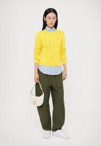 Κίτρινο πουλόβερ με πλέξη καλωδίου, ανοιχτόγαλάζιο πουκάμισο με γιακά από κάτω, χακί παντελόνι με φαρδιά πόδια, λευκά αθλητικά παπούτσια και μια μικρή μπεζ τσάντα χειρός.