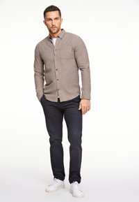 Camicia a maniche lunghe marrone con bottoni e tasca, abbinata a pantaloni blu navy e sneakers bianche. Design semplice con vestibilità casual e texture.