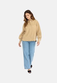 Beżowa koszula z korduroju na guziki z dwoma kieszeniami, połączona z jasnoniebieskimi spodniami z szerokimi nogawkami i czarnymi sandałkami na obcasie. Modelka spaceruje.