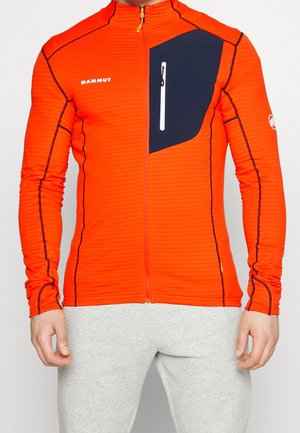 Mann trägt eine leuchtend orangefarbene Jacke mit durchgehendem Reißverschluss, schwarzer Brusttasche und schwarzer Naht, kombiniert mit hellgrauer Hose, Vorderansicht.