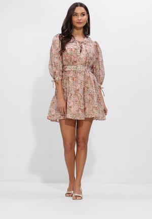 Donna in piedi che indossa un mini abito rosa a fiori con maniche a sbuffo, cintura e tacchi aperti, su uno sfondo bianco uniforme.