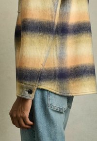 Camisa de franela con un patrón a rayas en tonos de naranja, amarillo y azul; textura suave, puños con botones, corte relajado, combinada con jeans de un azul claro.