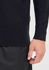 Svart genser laget av mykt materiale, med ribbede mansjetter og hem. Et uformelt merke er synlig på siden. Svart bukse under.