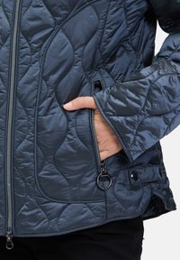 Gequilted marineblauwe jas met een glanzende afwerking, voorzien van een zilveren rits, zijzakken en een lussenvormig hardwareaccent aan de rits. 