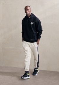 Felpa nera con cappuccio, tasca frontale e logo, abbinata a pantaloni jogger crema con strisce laterali nere e sneakers nere con dettagli verdi.