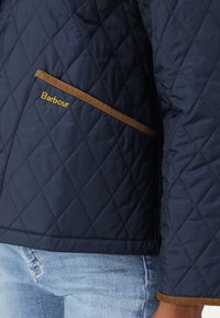 Granatowa pikowana kurtka z brązowym wykończeniem kieszeni, z złotym logo "Barbour". Widoczna teksturowana tkanina i dopasowany krój.