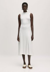 Witte, mouwloze jurk met een aansluitende lijfje, lichte flair aan de zoom en een gladde textuur. Gepaard met pale platte sandalen.