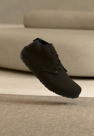 Zwarte veterschoen zwevend boven een beige tapijt met een lichtbeige bank wazig op de achtergrond.