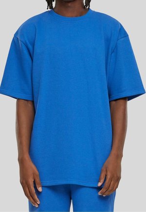 Person iført en ensfarvet blå oversized t-shirt med korte ærmer og matchende blå bukser mod en lys grå baggrund.
