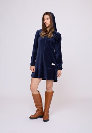 Marineblauwe fluwelen jurk met een capuchon, lange mouwen en een stikseldetail. Gecombineerd met hoge bruine leren laarzen met een laag hak.
