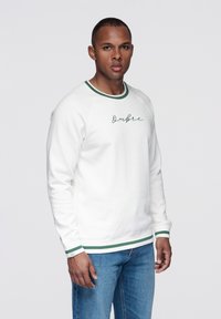 Vit sweatshirt med gröna detaljer vid kragen och ärmsluten, med kursiv text "Ombree", bärs med blå jeans.