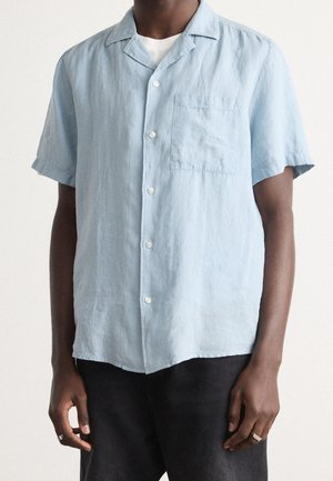 Lichtblauw overhemd met korte mouwen en knopen, met voorzak, gedragen over een wit ondershirt, gecombineerd met een donkere broek en zilveren ringen aan beide handen.