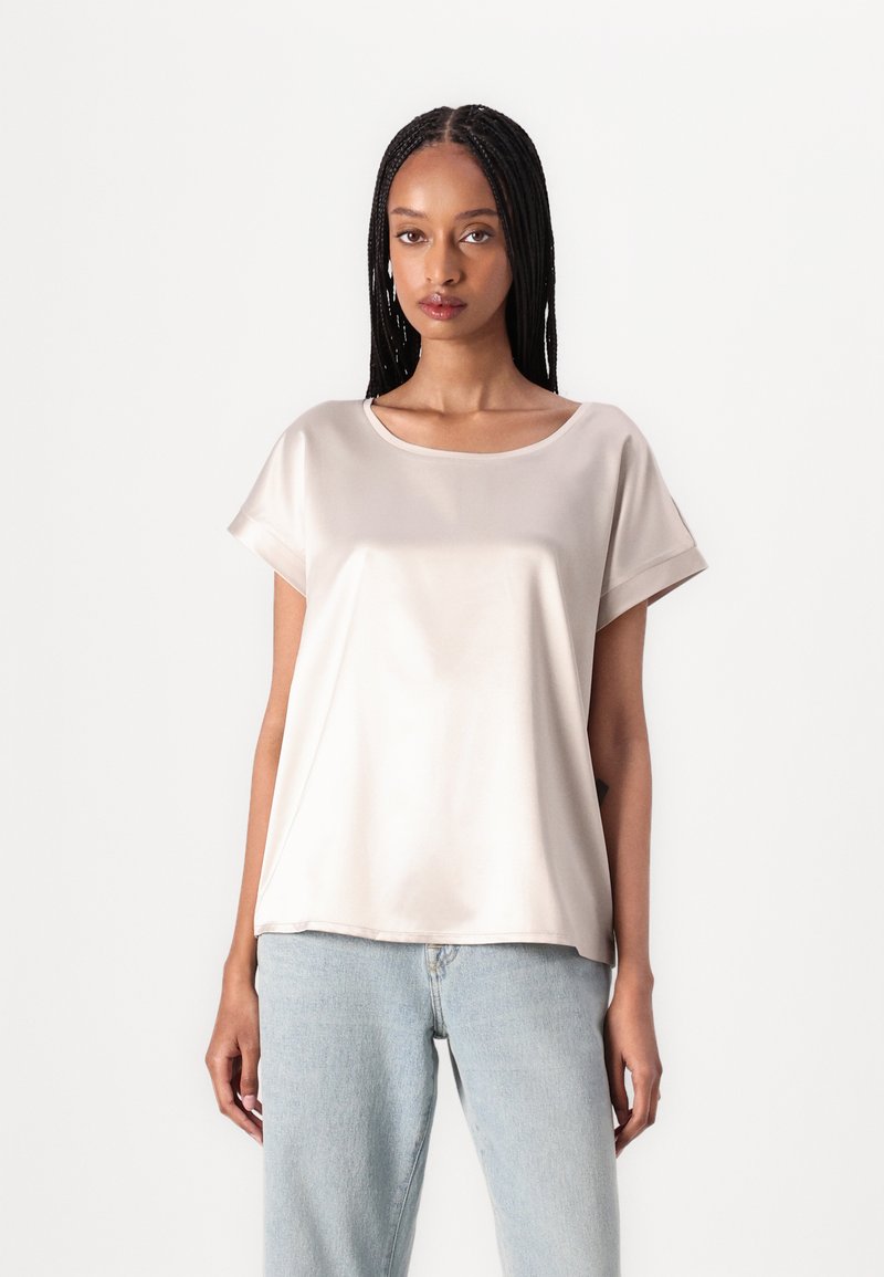 Satin kort mouwtop in lichtbeige met een ronde halslijn. De stof heeft een gladde, glanzende textuur. Gepaard met lichtblauwe denimjeans.