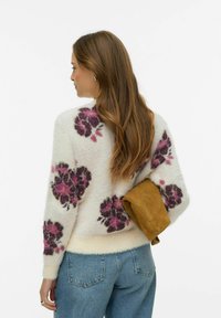 Balts pūkains džemperis ar ziedu rakstu, kurā ir violetas un rozā puķes. Modelis tur rokā brūnu zamša jaku un gaiši zilas džinsa bikses, skatīts no aizmugures.