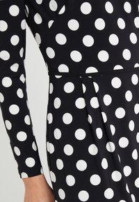 Svart klänning med vita prickar, långa ärmar, plisserad kjol och detaljer i midjan. Tyget verkar slätt och elastiskt.